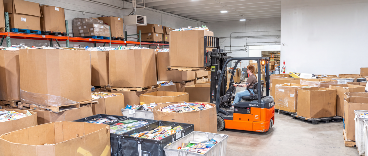 Pallets and boxes of used books, CDs and DVDs stacked in the BulkDiscs warehouse, with a forklift operator moving a pallet